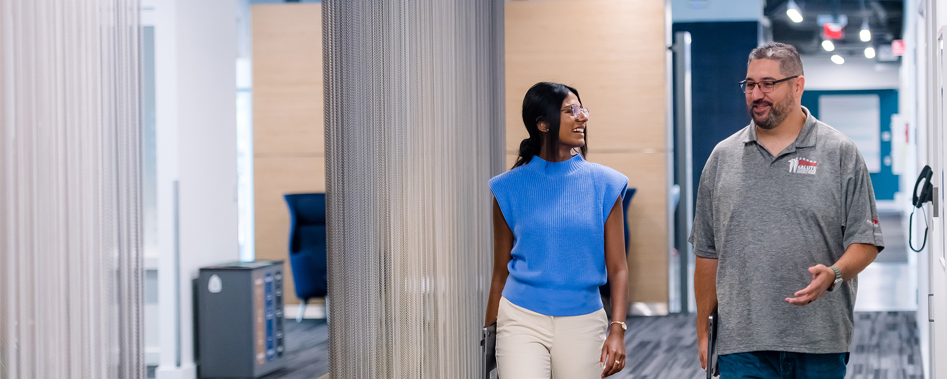 Two Capital One Tech Incubator associates walk down the hall in a Capital One building holding their laptops and talking