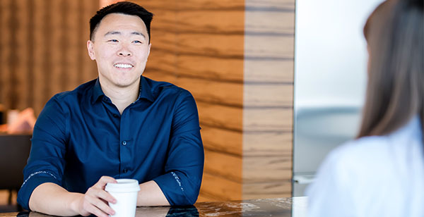 A Capital One Strategy Consulting associate sits across a table talking to another associate holding a cup of coffee wearing a blue collared shirt 