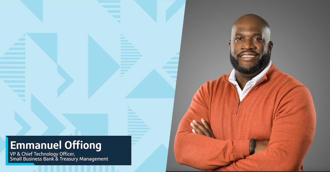 Capital One leader Emmanuel smiles in front of a gray background wearing an orange sweater