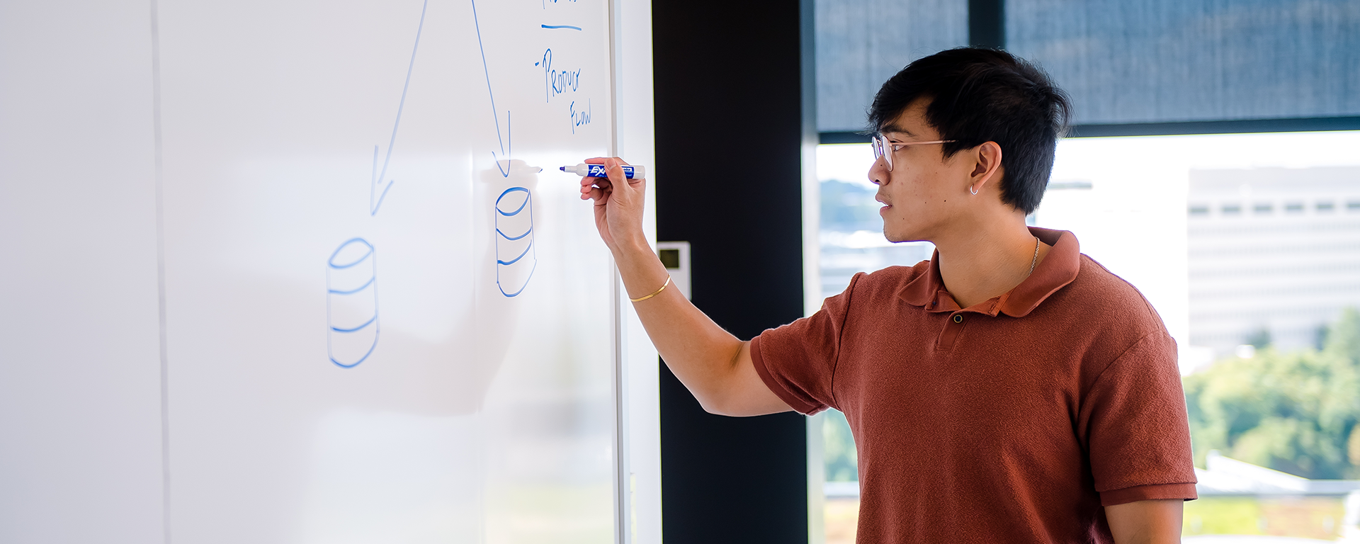 Capital One PDP Students & Grads associate writes on a whiteboard wearing a red shirt and glasses